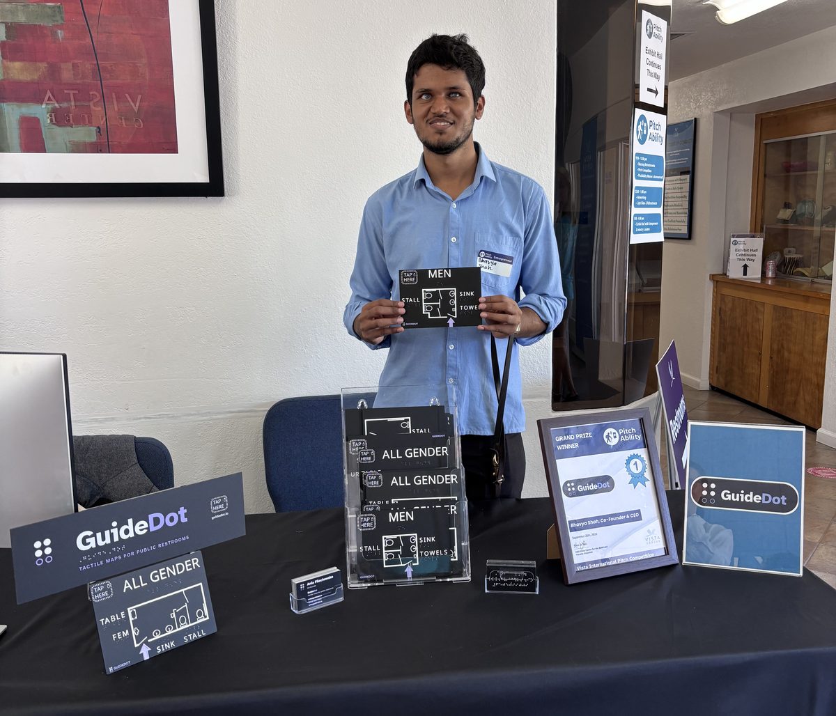 Bhavya smiles behind the GuideDot table at Vista Center in San Jose, holding up a small black MEN map with a TAP PHONE pad, white raised linework and the labels STALL, SINK and TOWELS. On the black tablecloth in front of him: a GuideDot sign, a stand of ALL GENDER and MEN maps, an ALL GENDER map on an easel, a braille business card, and a framed Grand Prize Winner certificate from the PitchAbility competition.