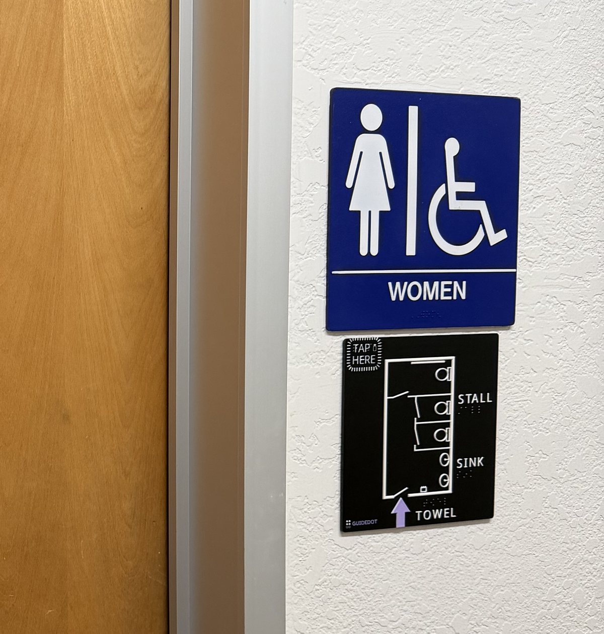 The edge of a wooden restroom door at Vista Center in San Jose, and on the white wall beside it the blue and white WOMEN sign with a wheelchair symbol. Directly below the sign hangs the GuideDot map: a small black plate with white raised linework showing the stalls, two sinks and a towel dispenser, the labels STALL, SINK and TOWEL in print and braille, a TAP HERE pad in the top corner, and a purple arrow at the doorway.