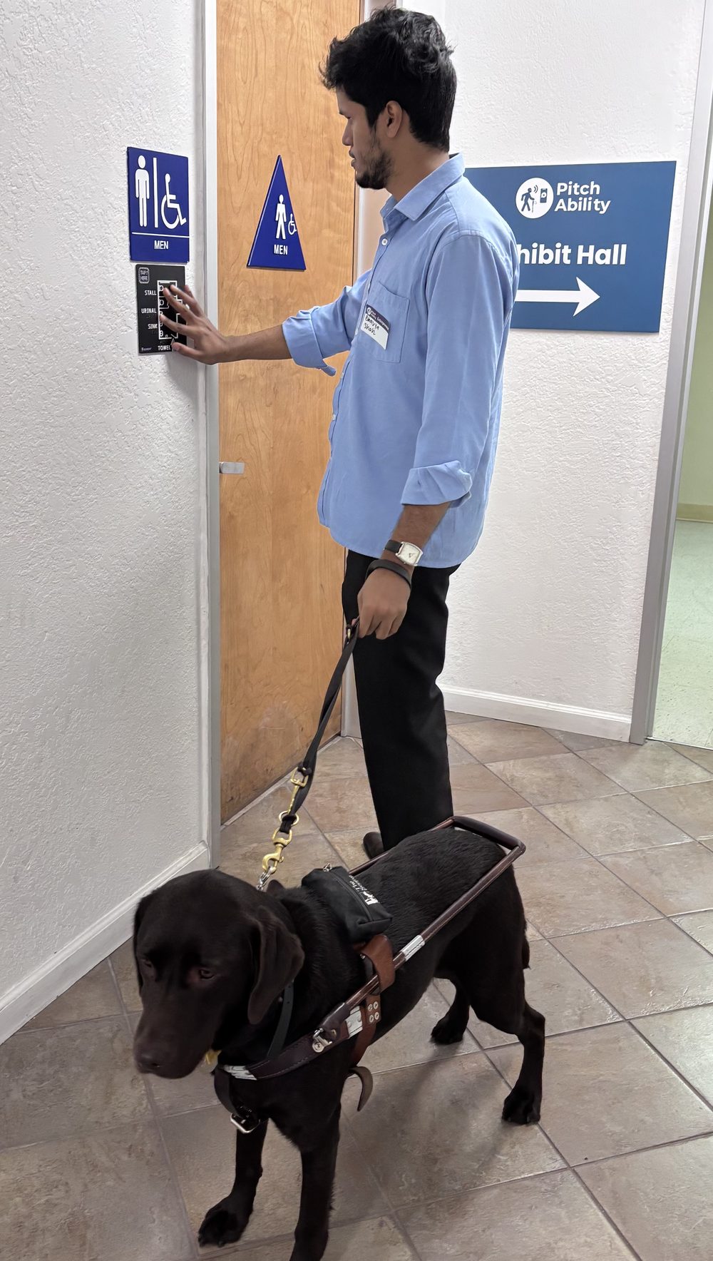 Bhavya, a young man in a light blue shirt, stands at the men&rsquo;s restroom door at Vista Center with his guide dog, a chocolate Labrador in harness, at his side. His right hand rests on the GuideDot map, a small black plate mounted directly below the blue and white MEN sign, with white raised linework, the labels STALL, URINAL, SINK and TOWEL, and a TAP HERE pad in the top corner.