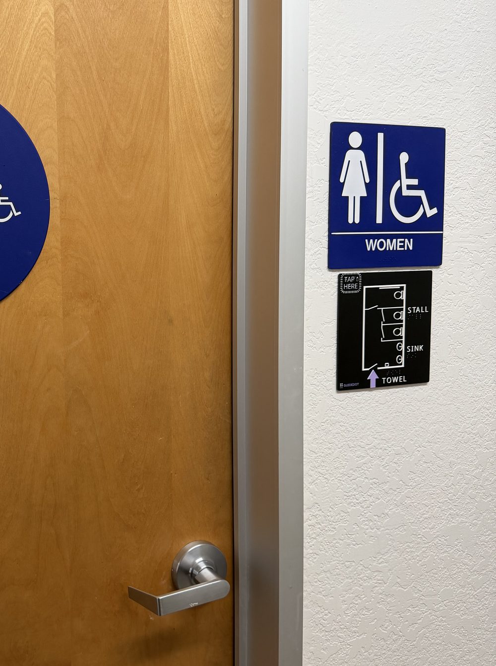 A wooden restroom door at Vista Center in San Jose with a round blue WOMEN sign on it. On the wall beside the door, the square blue and white WOMEN accessibility sign, and directly below it the GuideDot map: a small black plate with white raised linework showing two stalls, a sink and a towel dispenser, the labels STALL, SINK and TOWEL in print and braille, a TAP HERE pad in the top corner, and an arrow at the doorway.